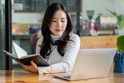 Young woman holds open book while looking at laptop screen