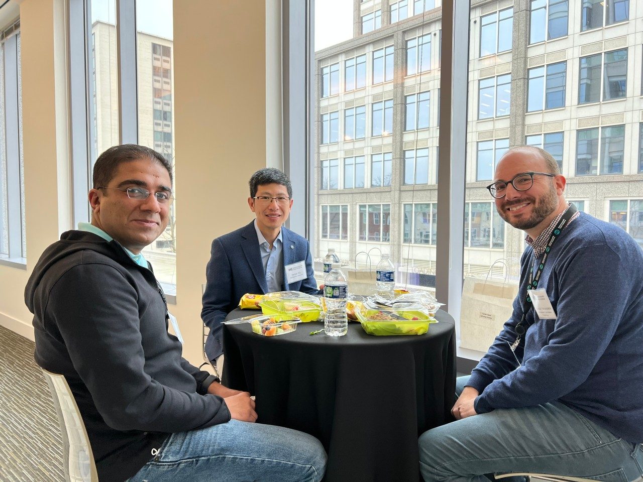 Three people sit around a small round table near large windows in a bright office space.