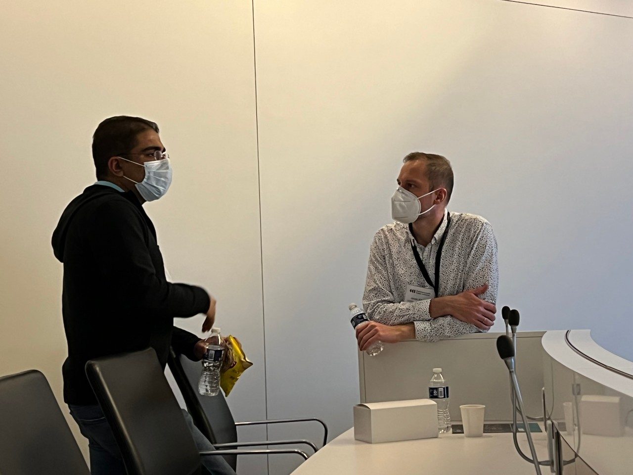 Two people talking in a conference room near a curved table with microphones. 