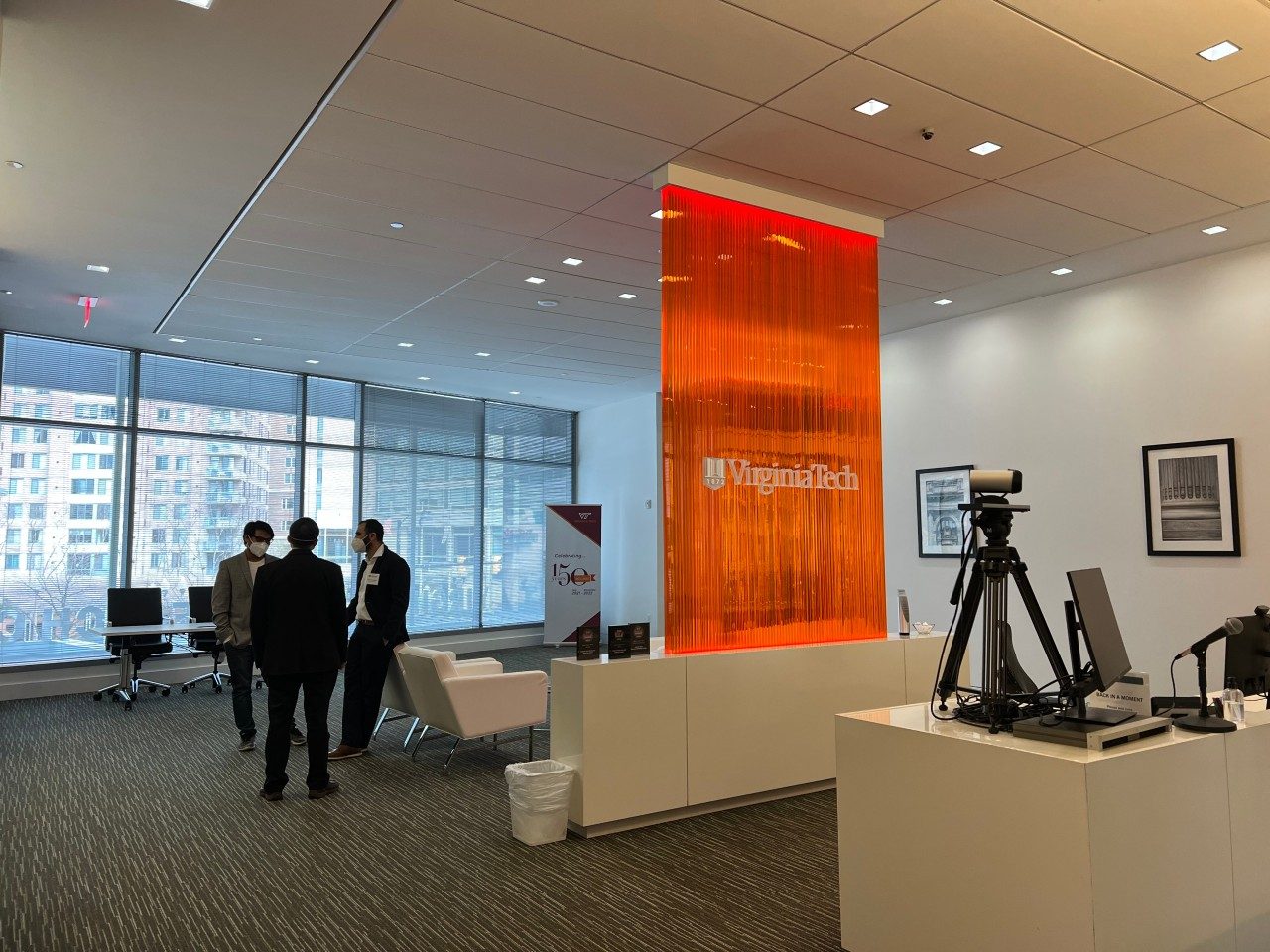 Three people stand near a seating area talking, while a desk in the foreground holds camera equipment and office supplies.