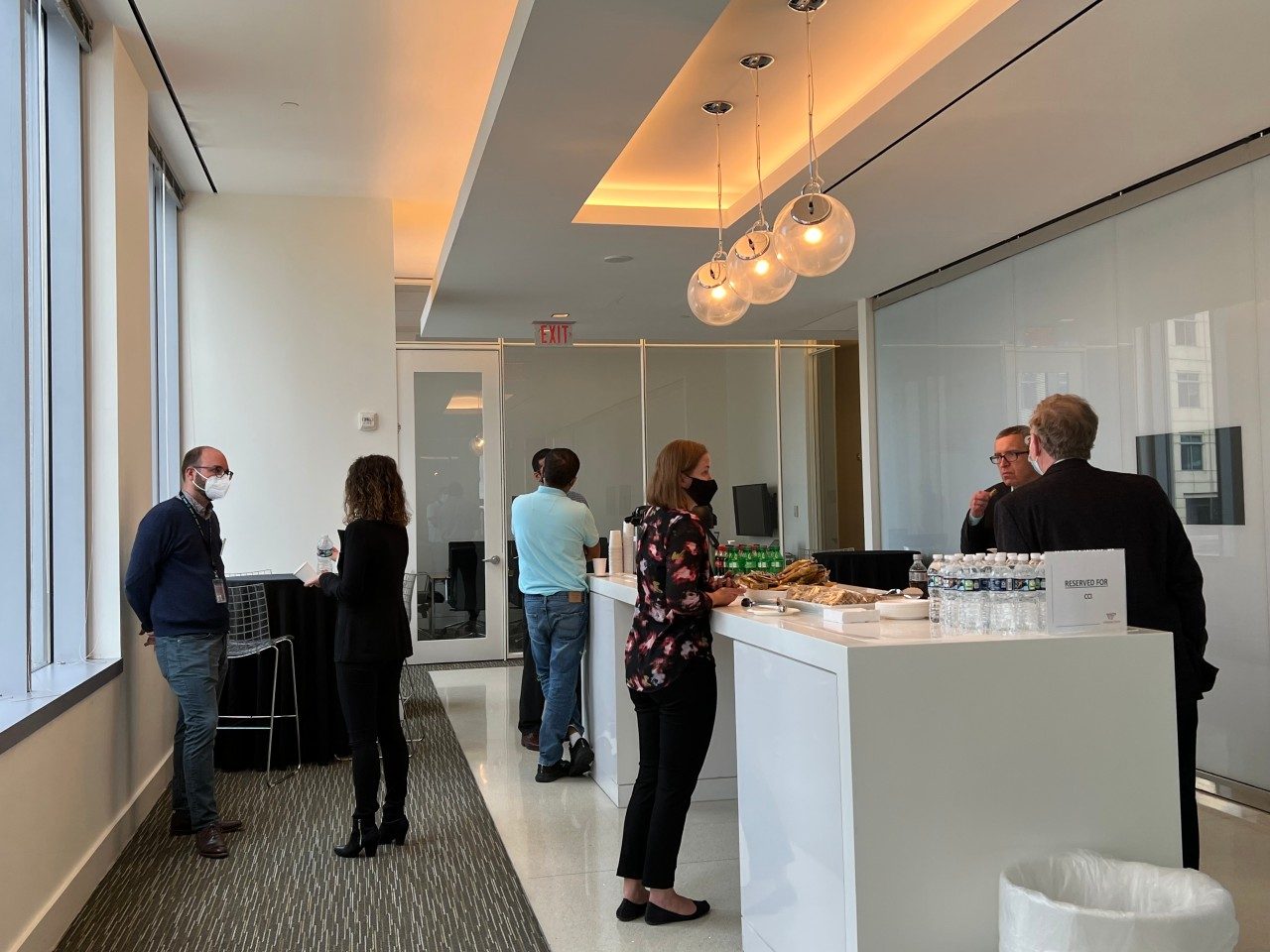 Several people gather in a bright office hallway near a counter with snacks and bottled drinks.