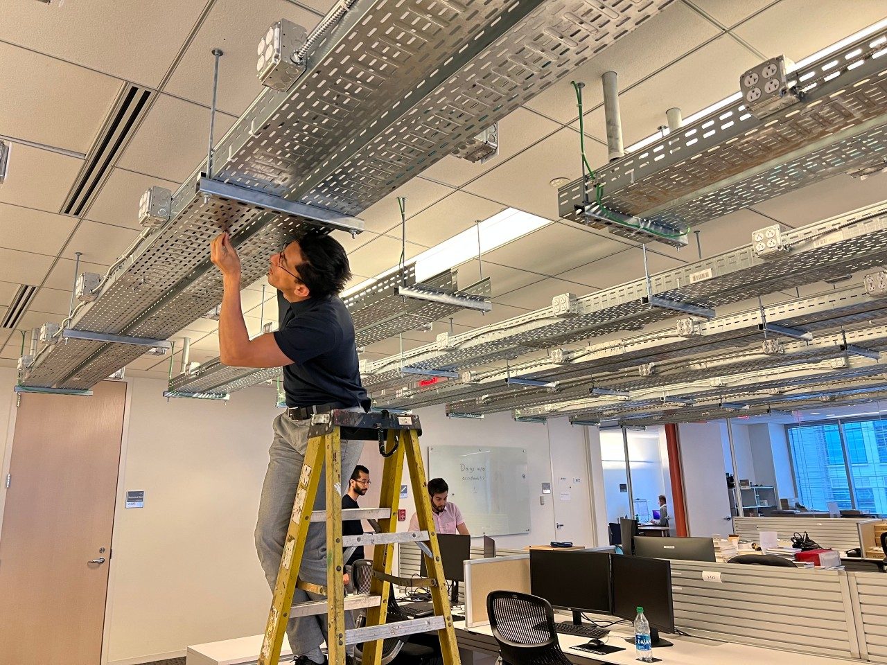 A person stands on a ladder in an office space working on overhead metal cable trays.