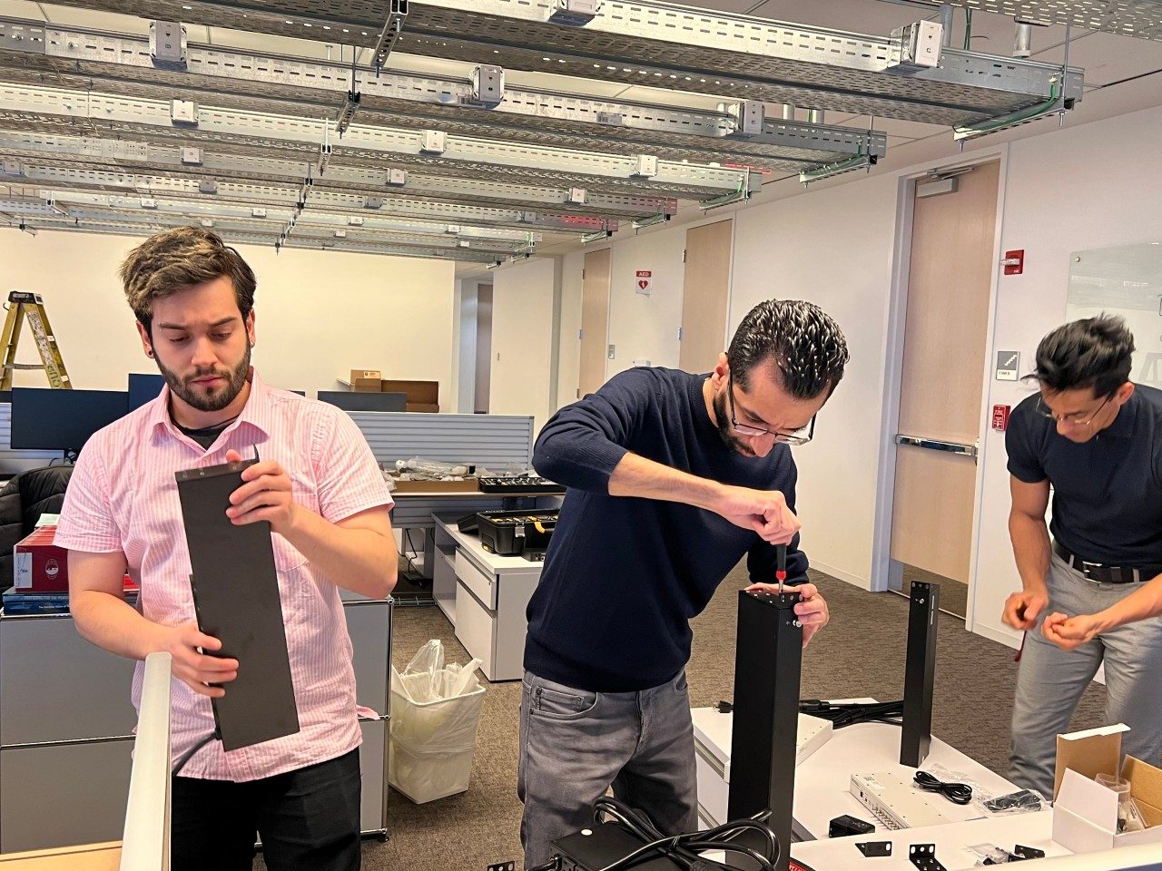 Three people work together in an office assembling equipment on a table.