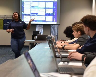 Student stands in front of screen as students on laptops watch