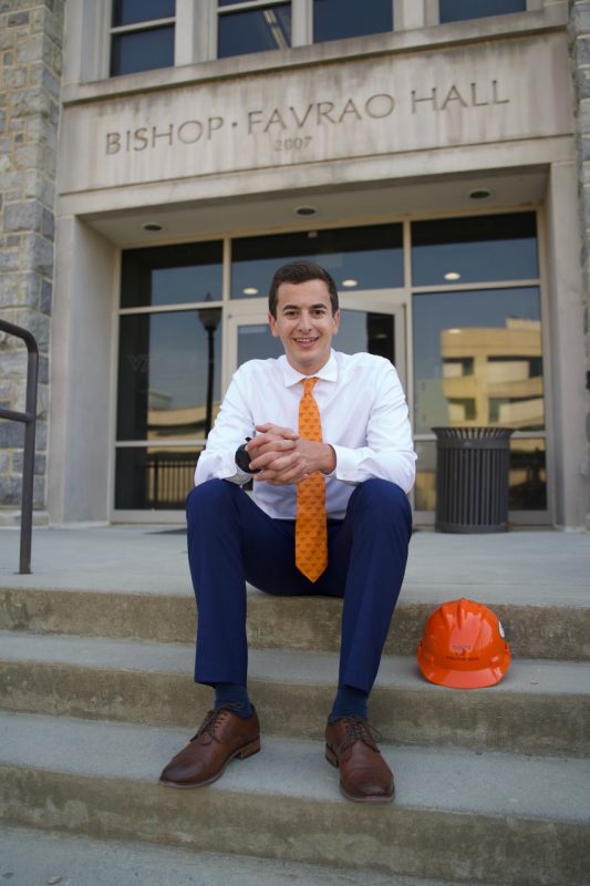 Man sits outside of academic building in a suit and Virginia Tech tie.