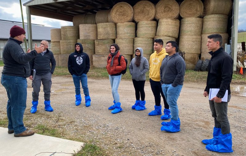 A teacher stands in front of a group of students who are all wearing blue, plastic boots.